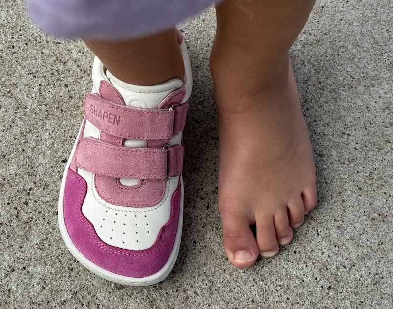 A top down view of a child's feet standing on concrete with one foot bare and the other foot wearing a kids Shapen Barefoot sneaker with a wide toe box, pink accents, and velcro straps.