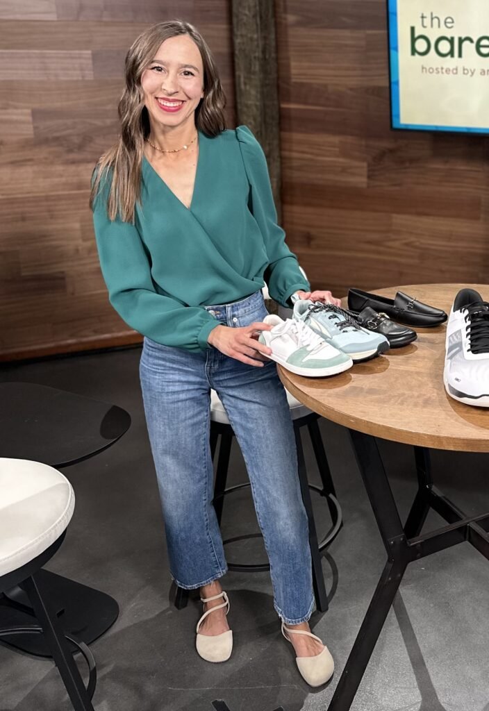 A woman standing next to a table holding shoes, smiling at the camera wearing a dressy blouse and Shapen Barefoot Orchid flats in beige suede leather
