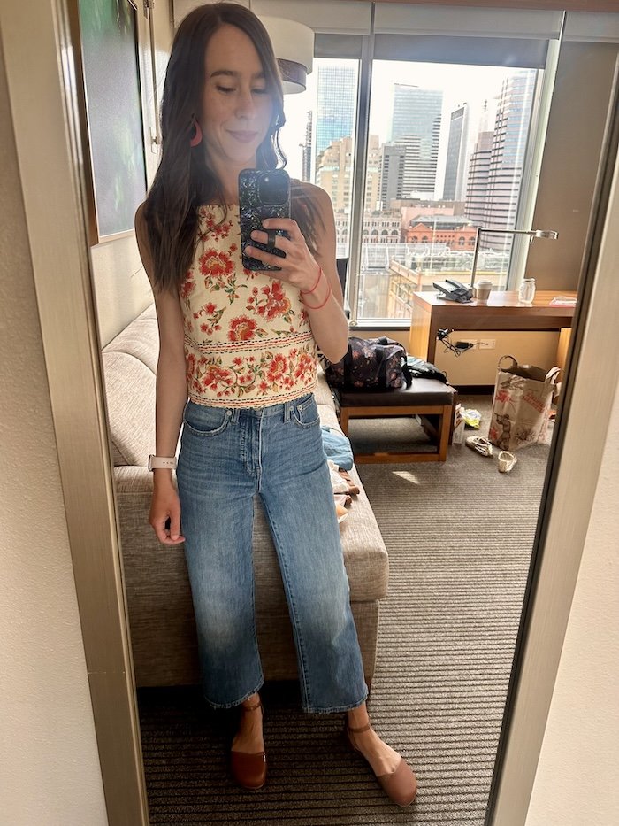 A woman taking a selfie in a mirror wearing jeans, a floral top, and Shapen Barefoot Poppy flats in cognac color, with a wide anatomical shape