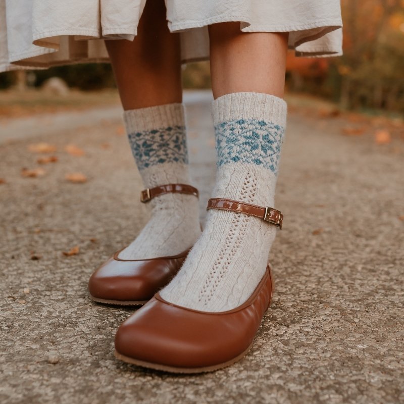 Close up of a pair of socked feet wearing Shapen Tulip flats in Cognac. Fallen leaves are blurred in the background and the person's natural colored dress is just visible at the top of the frame. The socks have dainty details and tiny blue snowflakes around the cuff.