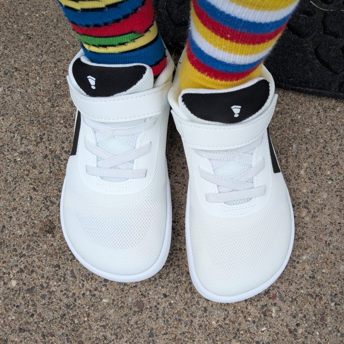 Top down view of Splay Dash white barefoot sneakers for kids with black accents. The wide natural shaped toe box is in the center of the image. Mismatched colorful socks are worn with the shoes.