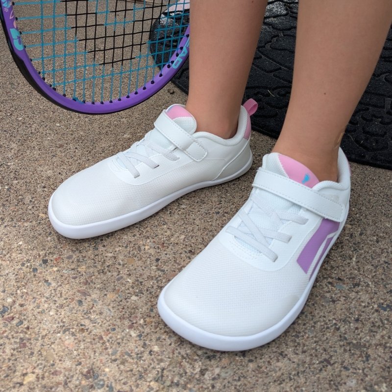 A close up side view of Splash Dash barefoot wide toe box sneakers for kids in white with purple and pink accents. A matching purple tennis racket is seen in the background