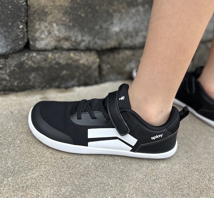 A close up side view of a child's foot standing on pavement wearing Splay Dash kids barefoot play shoes in black