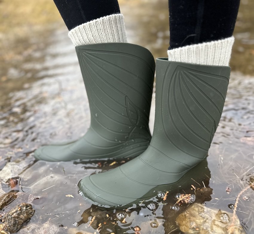 A side view of a person's lower legs wearing the Wildling Ranidae barefoot wide toe box rubber rain boots in olive green standing in a puddle with wool socks poking out the top