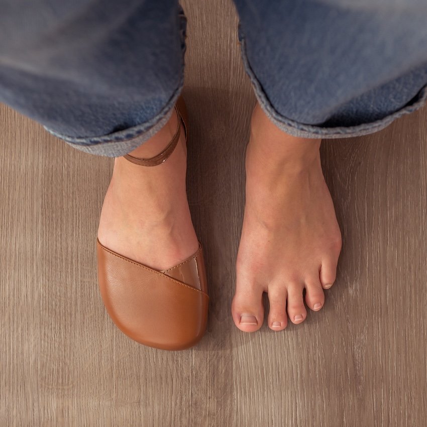 A top down view of a pair of feet standing on a wood floor. The right foot is bare and the other foot is wearing a Shapen Barefoot Poppy in Cognac leather, wide width.