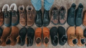 Two lines of barefoot boots placed toe to toe, with one person's feet in a pair of Peerko black lace up barefoot boots. A variety of brands are represented including Shapen, Be Lenka, Mukishoes, and Zaqq.