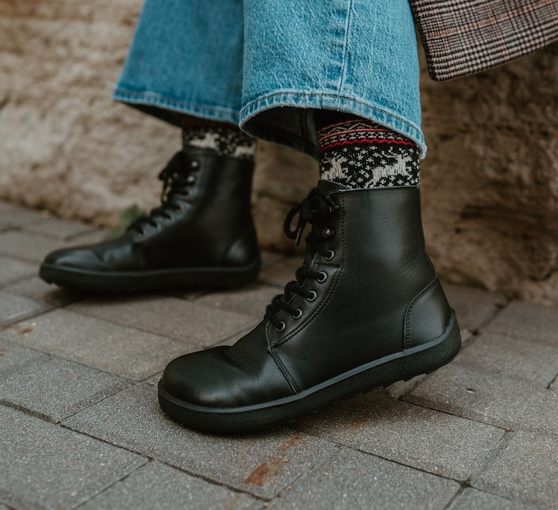 A close up side view of a pair of black combat leather lace up winter boots from Be Lenka being worn by a person sitting on a stone wall. A plaid jacket is just visible at the top of the frame, and wide leg denim are resting above the boots with a pair of chunky knit socks peeking out over the boots.