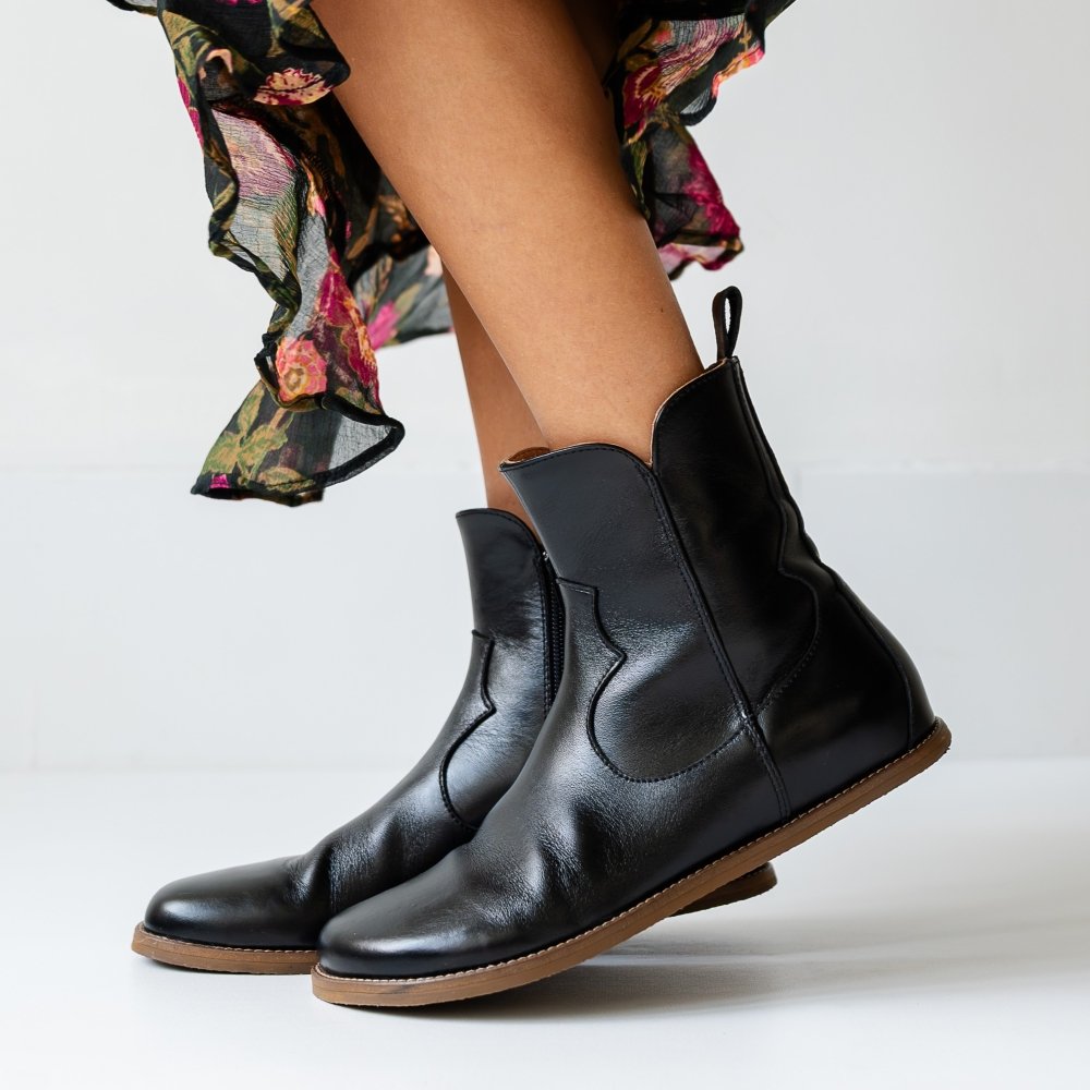 Side view of a pair of Faye Boots in black leather worn with a pretty floral skirt. The front foot is lifted slightly to show off the zero drop boot.
