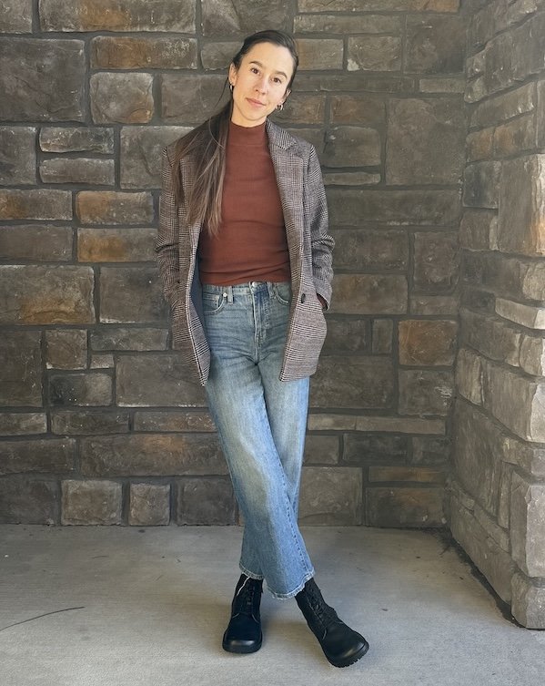 A woman standing against a stone background with her legs crossed and her hands in her plaid jacket pocket. She has on a rust colored top and black lace up boots from Lang S Tikki Shoes, a sustainable barefoot shoe brand. The black leather Kombat boots give the outfit a bold finish.