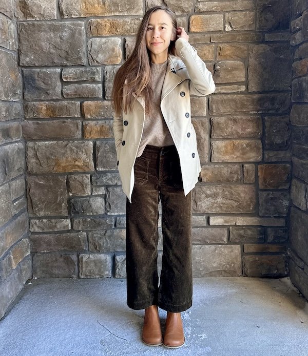 Full body view of a woman standing in a stone alcove wearing cropped wide leg dark brown corduroy pants, a khaki jacket, and sustainable barefoot wide toe box boots from Lang S Tikki Shoes in a light brown leather.