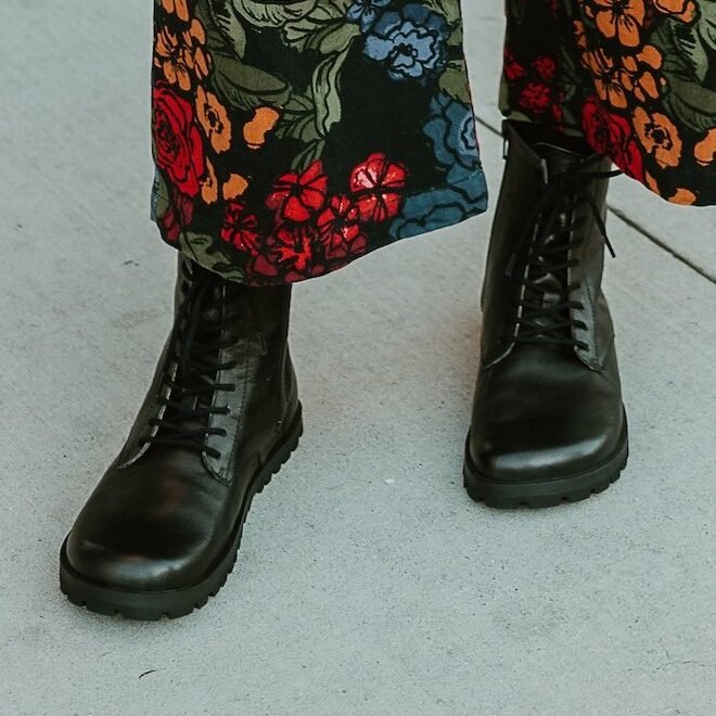 Cropped look at Lang S Kombat black leather barefoot boots being worn by a person in a floral jumpsuit. The boots show off a wide comfortable toe box and a flat, flexible outsole.