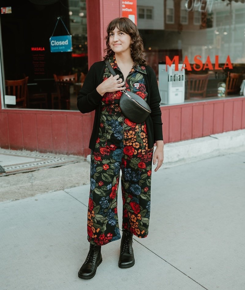 Full body view of a woman with wavy brown hair standing on a downtown sidewalk wearing a floral jumpsuit, black sweater, and black bag. She is looking off into the distance with a slight smile on her face. The outfit is finished with Lang S Kombat black leather lace-up barefoot boots.