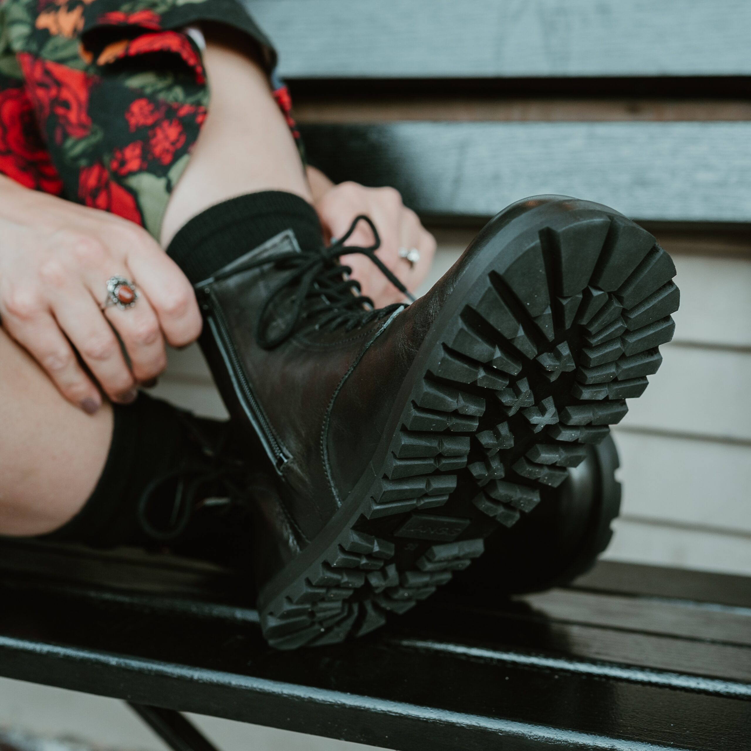 A zoomed in view of a woman sitting on a bench with her Lang S Kombat boots propped up in front of her adjusting the laces. The chunky zero drop outsole is in the forefront of the frame, and a flowery jumpsuit is blurred in the background. An inside easy access zip is also shown, along with adjustable laces for a classic look.