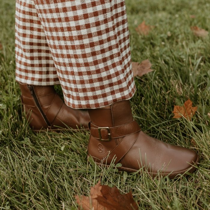 A person standing on a leafy ground wearing a pair of gingham pants with coordinating Shapen Divine brown barefoot boots with buckle strap details