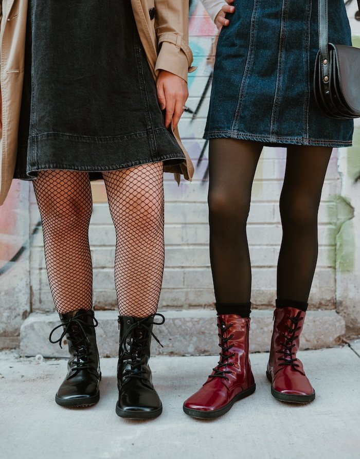 Two women standing next to each other in a graffitied alleyway wearing denim mini dresses. On the right, she is wearing maroon red patent leather lace up Maverick boots by Shapen Barefoot, and on the left, a matching pair in black patent leather with fishnet tights.