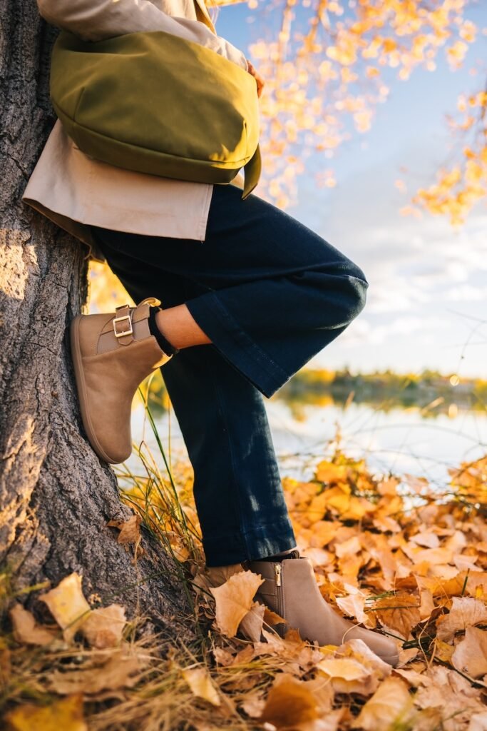 A chest down view of a woman leaning against a tree with fall yellow and brown leaves scattered on the ground. She has a green leather bag resting on her waist, and her front foot is propped up on the tree trunk. On her feet are a pair of taupe leather barefoot chelsea boots from Xero Shoes. In collaboration with Anya, the Tari II worn here has an inside zipper and outside buckle detail with elastic. Wide leg dark denim are being worn by the model, along with a khaki jacket.