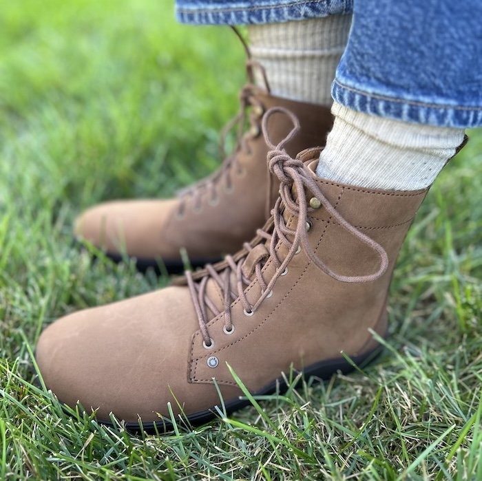 A close up front side view of Xero Shoes barefoot Breckenridge lace up boots in brown leather with a wie toe box and flexible outsole. A pair of mid-tone denim is visible at the top of the frame, bright green grass is underfoot, and a pair of slouchy cream socks are filling the space between the boot and jeans.
