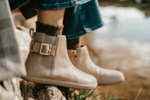 Close up detailed view of a pair of taupe barefoot chelsea boots from Xero Shoes. The elastic and buckle detail are in the forefront of the image, and the inside zipper is just visible on the boot in the background. The zero drop outsole is also featured. A pair of chunky brown socks are being worn and are peeking out above the boot.