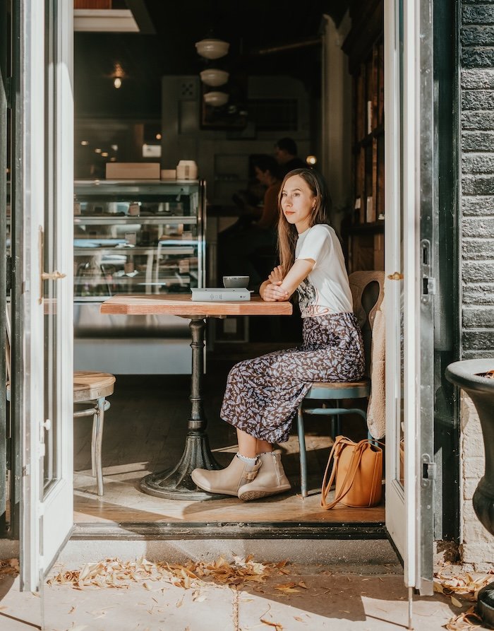 A photo looking through an open cafe door at a woman sitting alone at a table. She has a closed book in front of her and her arms are crossed resting on the table. She's looking off into the distance with a slight smile on her face. She is wearing a floral skirt and a graphic t-shirt pair with a pair of taupe ankle boots with a buckle and wide toe box from Xero Shoes.