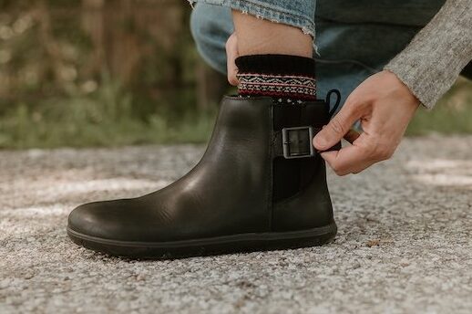 A close up view of a person in Tari II black leather buckle anke barefoot boots by Xero Shoes and Anya crouched down adjusting the buckle on one boot. The boot in back in flexed and the camera is focused on the front boot, which has been paired with a patterned sock.