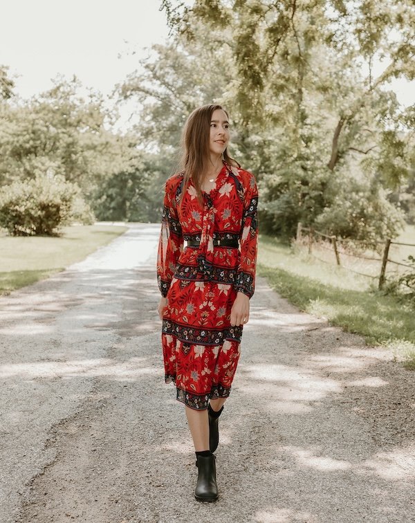 A woman walking down a shady road looking off to the side with a red patterned dress, paired with a pair of black barefoot chelsea boots from Xero Shoes with a ankle buckle and zipper detail. The Tari II boot featured a wide anatomical toe box and flexible outsole for foot health.
