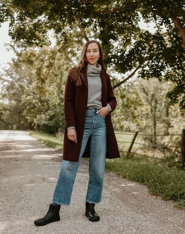 A woman standing outside smiling at the camera wearing jeans, a burgundy coat, and black Xero Shoes Tari II barefoot chelsea boots designed by Anya from Anya's Reviews