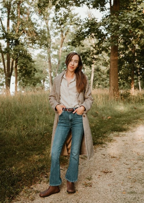 A woman standing near the edge of the a wooded area, wearing flare leg jeans, a cream button up shirt, and a long herringbone wool coat. Her hands are in her pockets and she is looking at the camera. Her right foot is turned out and she has brown leather Xero Shoes Tari II boots on her feet, a wide toe box chelsea boot alternative.