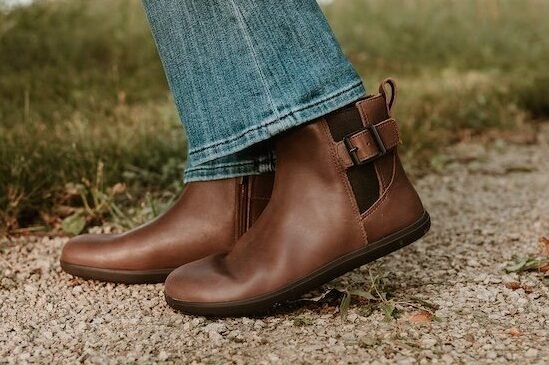Close up side profile view of a pair of dark brown buckle boots, the Tari II, a collaboration between Anya and Xero Shoes. The shoe in the foreground is lifted slightly showing off the zero drop flexible outsole, and ankle buckle. The back foot is flat on the ground and the inside zip is just visible. A pair of flared leg jeans are resting just above the boot opening.