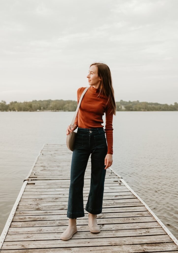 A woman in cropped dark denim and a burnt orange long sleeve top, standing on the dock of a lake with a handbag over her shoulder. She is looking off to the left with a slight smile on her face. On her feet are a pair of barefoot chelsea boots from Xero Shoes, a collabration with Anya's Reviews. The wide toe box taupe Tari II finishes the woman's outfit with a quiet elegance.