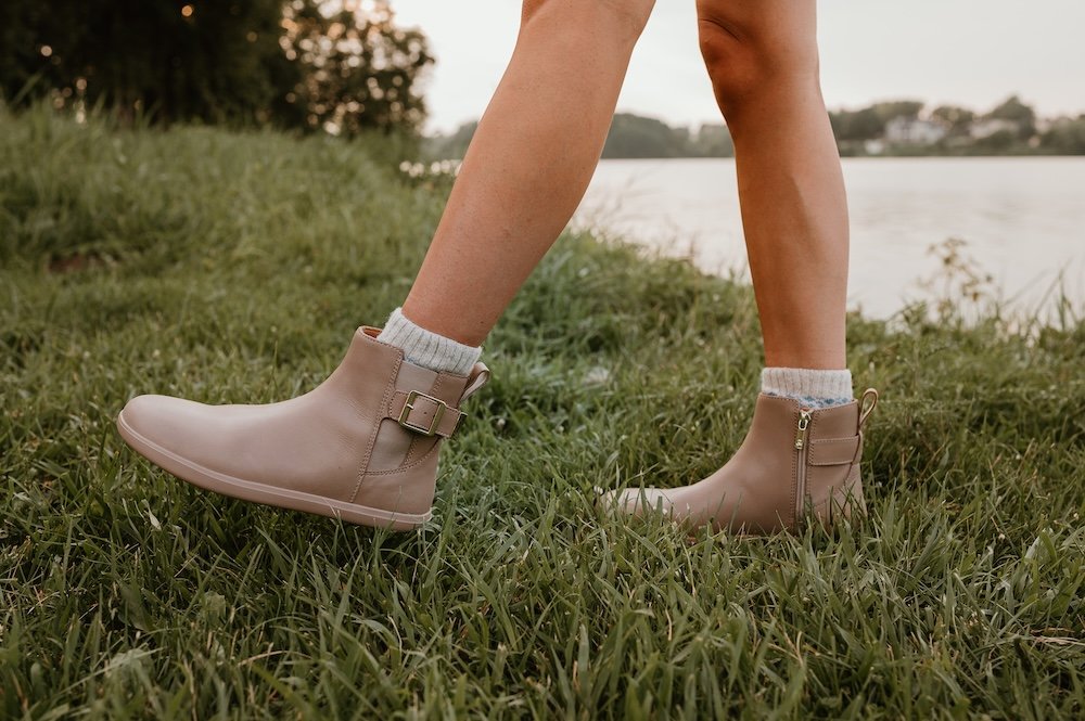 A woman's legs visible from the knees down standing on grass with one foot kicked out in front, wearing cozy knit socks and taupe doeskin Xero Shoes Tari II barefoot chelsea boot co-designed with Anya from Anya's Reviews