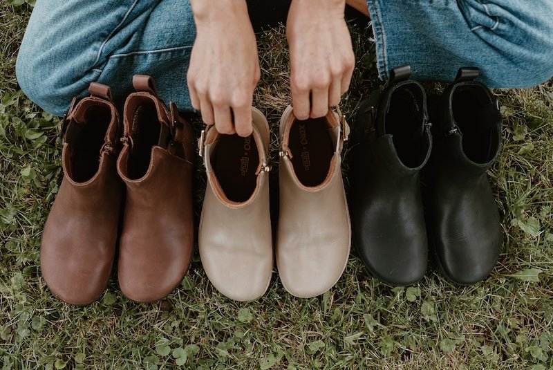 A line of Tari II barefoot chelsea boots in 3 leather colors - brown, taupe, and black. A woman is sitting cross-legged behind the line-up and is holding the back of the boots. All 3 pairs show the wide, anatomical toe box shape.