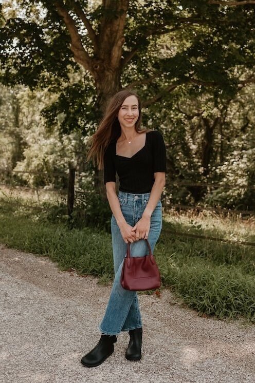 Full body photo of a woman with long brown hair standing with her legs crossed in front of her, holding a red handbag, and smiling at the camera. She is photographed outside under a shady tree on a gravel path, her black top matches her black leather barefoot boots - the Tari II from Xero Shoes.