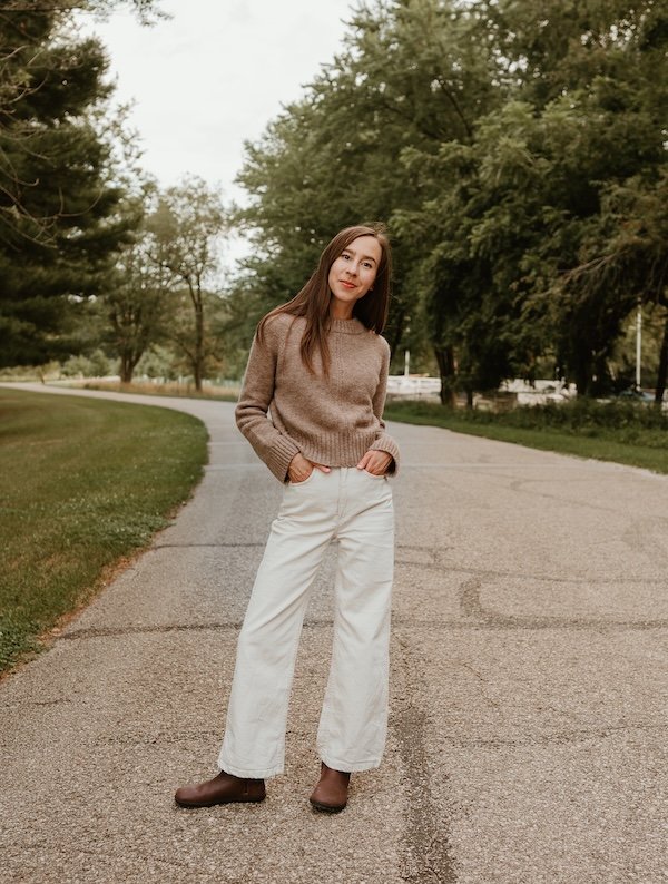 Full body view of a woman in wide leg cream colored jeans and a light brown sweater standing in a narrow road with her hands in her pockets and one leg turned out. On her feet are a pair of brown leather Tari II barefoot chelsea boots from Xero Shoes. The wide toe box and zero drop outsole provide all day comfort.