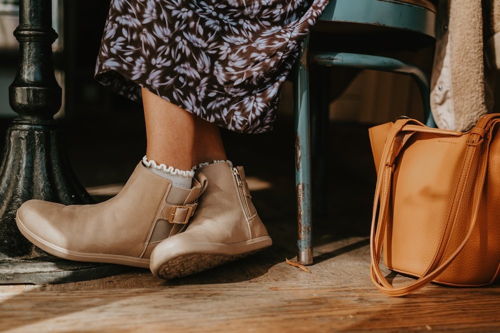 A close up of a woman's feet and lower legs crossed at the ankles. She is wearing white socks and Xero Shoes Tari II x Anya doeskin chelsea boots
