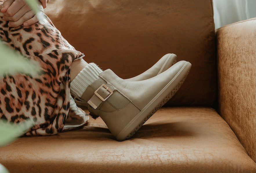 A pair of legs visible from the knee down resting on a brown couch. The person is wearing a leopard print skirt and Xero Shoes doeskin taupe barefoot Tari II chelsea boots, designed with Anya from Anya's Reviews