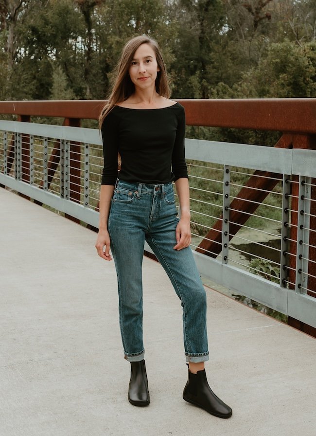 A woman standing on a bridge wearing denim, a black shirt and Zaqq Barefoot Equity chelsea boots in black leather