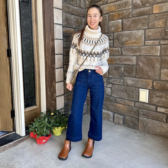 A woman standing on her front porch wearing dark cropped wide leg denim, a cozy sweater, and Feelgrounds Chelsea barefoot vegan boots with a wide toe box.