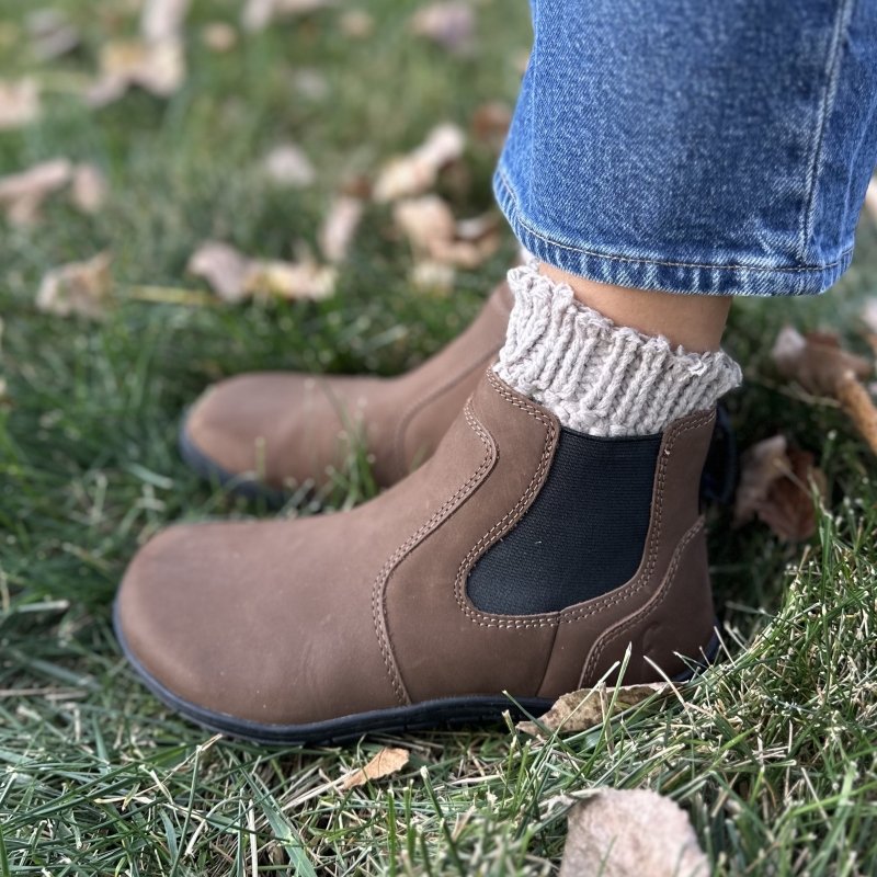 Side profile view of a pair of zero drop barefoot Freet Durham chelsea boots showing the detailed seams and side elastic. The person is standing on leaf strewn ground and chunky knit socks are peeking out the top of the boots.