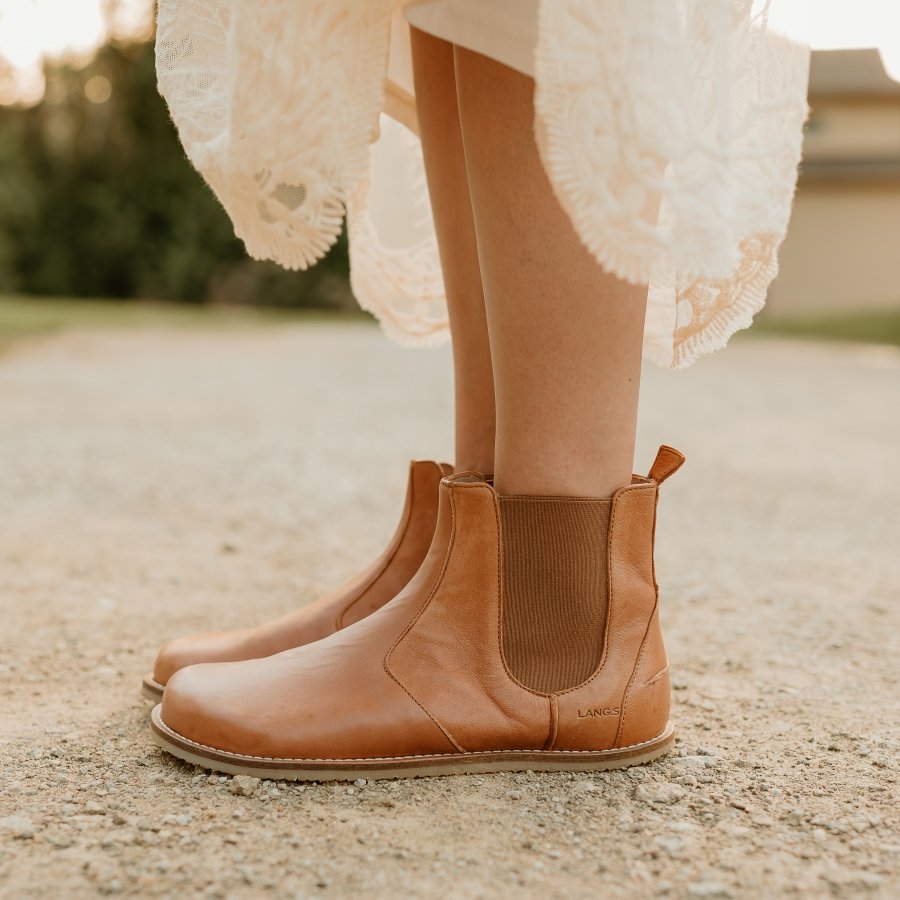 Side profile of a woman in Lang S chelsea boots in a caramel leather color with a lace dress at the top of the frame. The zero drop flexible outsole provides ground feel against the gravel road.