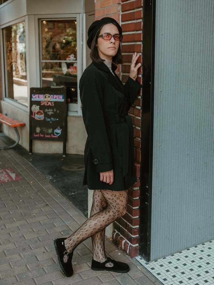 Full body view of a woman in a black trench coat, sunglasses, black hat, and fishnet lace tights. Her outfit is finished with a pair of Lang.S T strap flats and she's leaning against a downtown brick building.