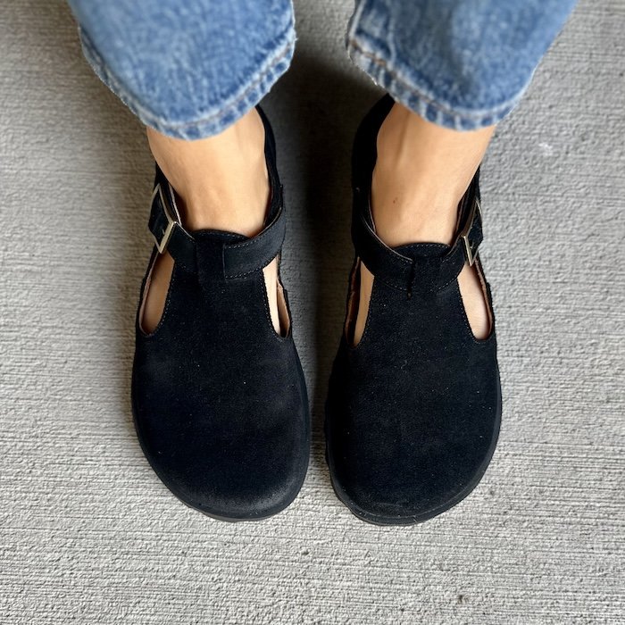 a top down view of a person's feet standing on concrete wearing jeans and Lang.S Mira black suede t-bar flats with a wide toe box