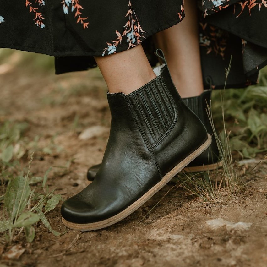 Side profile view of a pair of Lila chelsea leather barefoot boots by Anya's Shop. A black floral skirt is visible at the top and a dirt path is underfoot. The leather covered elastic is showing on the outside of the boot.