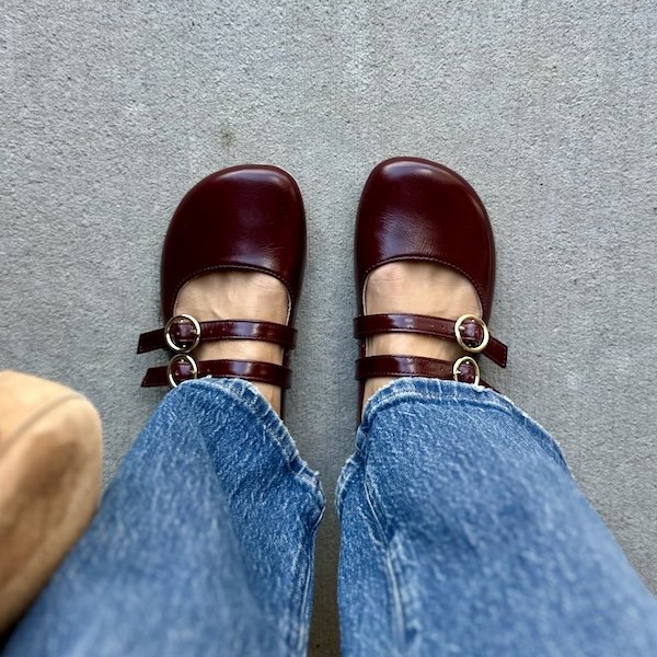 A top down view of a pair of women's feet wearing long denim and Origo Shoes barefoot two strap mary janes in patent burgundy leather