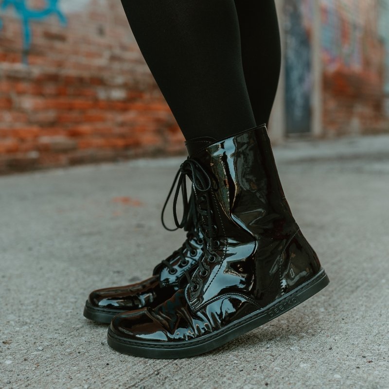 Side view of Peerko pulse patent leather combat style barefoot boots shown from the side. A brick graffitied wall is blurry in the background