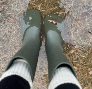 A top down view of a person wearing pair of 100% rubber rain boots with a wide toe box and a felt lining. Boots are green, and the person is wearing wool socks with them and standing in a puddle of water a couple of inches deep