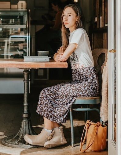 A photo looking through an open cafe door at a woman sitting alone at a table. She has a closed book in front of her and her arms are crossed resting on the table. She's looking off into the distance with a slight smile on her face. She is wearing a floral skirt and a graphic t-shirt pair with a pair of taupe ankle boots with a buckle and wide toe box from Xero Shoes.