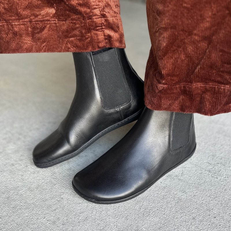 Side view of a pair of feet in Zaqq Equity barefoot leather chelsea boots with no seam details, a flat flexible outsole, and anatomical to ebox. A pair of crinkly burnt orange wide leg corduroy pants are resting above the boot