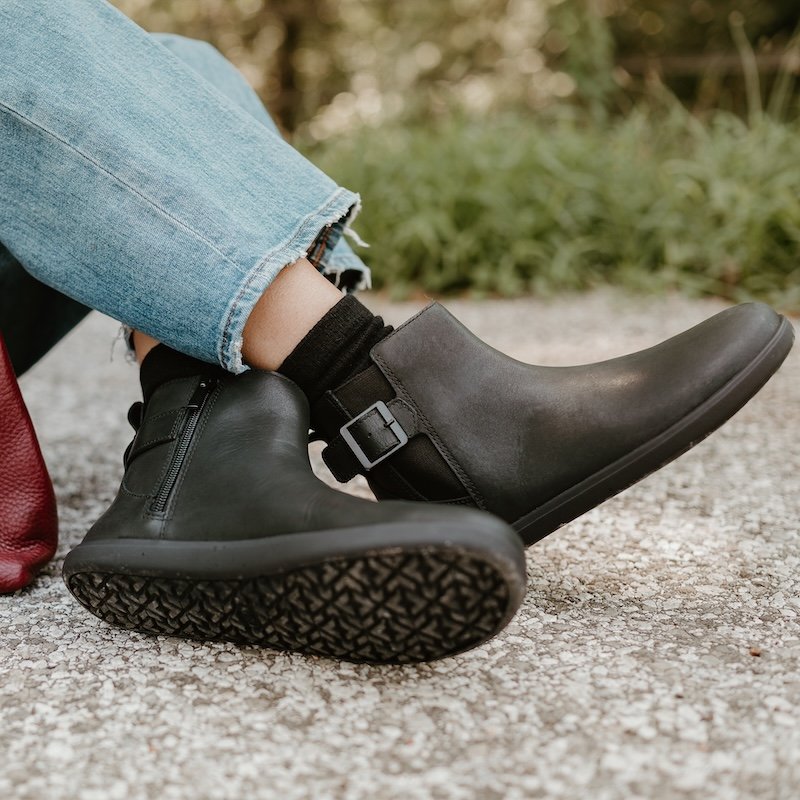 A close up of a pair of crossed feet against a rocky path. The person is wearing black leather barefoot Xero Shoes Tari II chelsea boots with a buckle detail. The textured outsole is visible at the bottom of the photo and tall grass is blurry in the background.