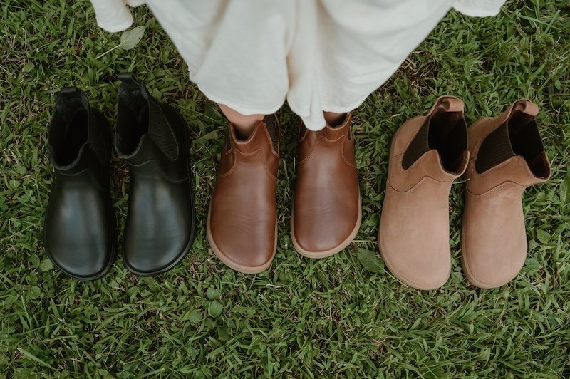 A line up of Be Lenka Entice barefoot extra wide toe box chelsea boots in 3 colors. Black on the left, dark brown in the middle worn by a woman in a long cream dress, and a light brown latte color on the right