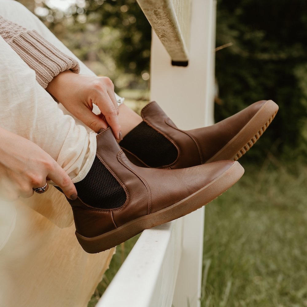 A close up of a woman's feet resting on a white picket fence. She is wearing a cream flowy dress, and a taupe sweater. Her hands are adjusting the brown leather Be Lenka Entice barefoot chelsea boots.
